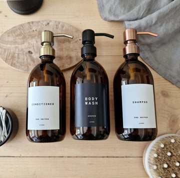three amber bottles with brass, matte black and copper metal pump and a personalised label in black and white version that says shampoo, body wash, conditioner. with the family name underneath for extra personalised touch