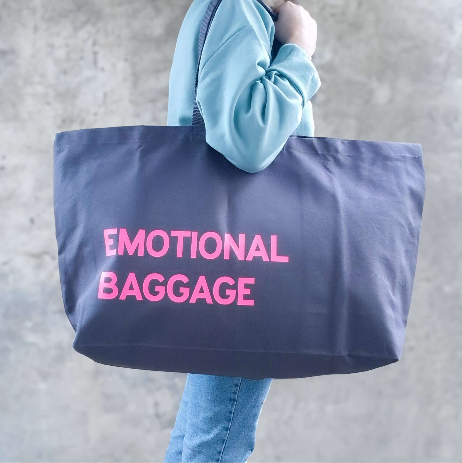 Grey oversized tote bag with neon pink text ‘emotional baggage’, displayed against a neutral background.