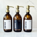 Three brown bottles with gold pumps labeled 'Hand Wash', 'Shampoo', and 'Hand Lotion' on a marble surface.