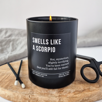 Black candle labeled 'Smells Like a Scorpio' on a wooden tray with a white background
