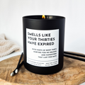 Black candle with a humorous label 'Smells like your thirties have expired' on a wooden surface
