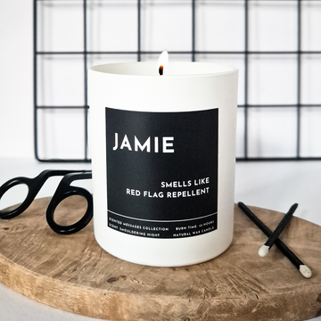 White candle with a black label and a text with personalised name and a text ' smells like a red flag repellent' on a wooden surface.