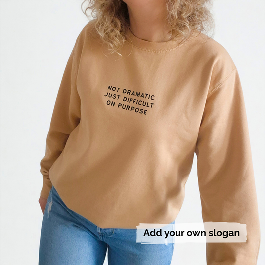 Person wearing a tan sweatshirt with text 'Not Dramatic just difficult on purpose' and blue jeans on a white background