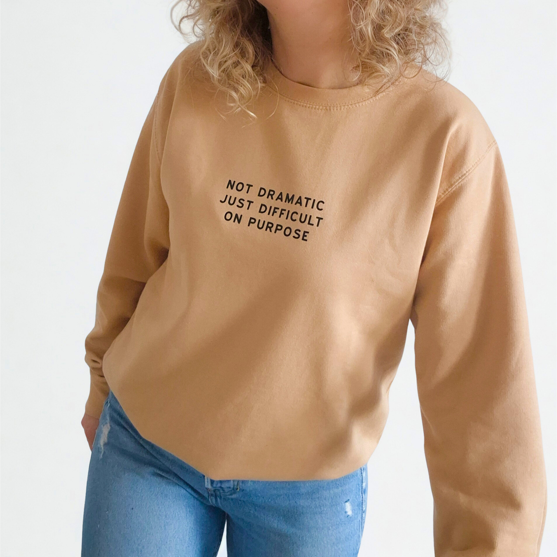 Person wearing a beige sweatshirt with text 'Not dramatic just difficult on purpose' and blue jeans on a white background