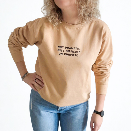 Person wearing a beige sweatshirt with text  'Not dramatic just difficult on purpose' and blue jeans on a white background