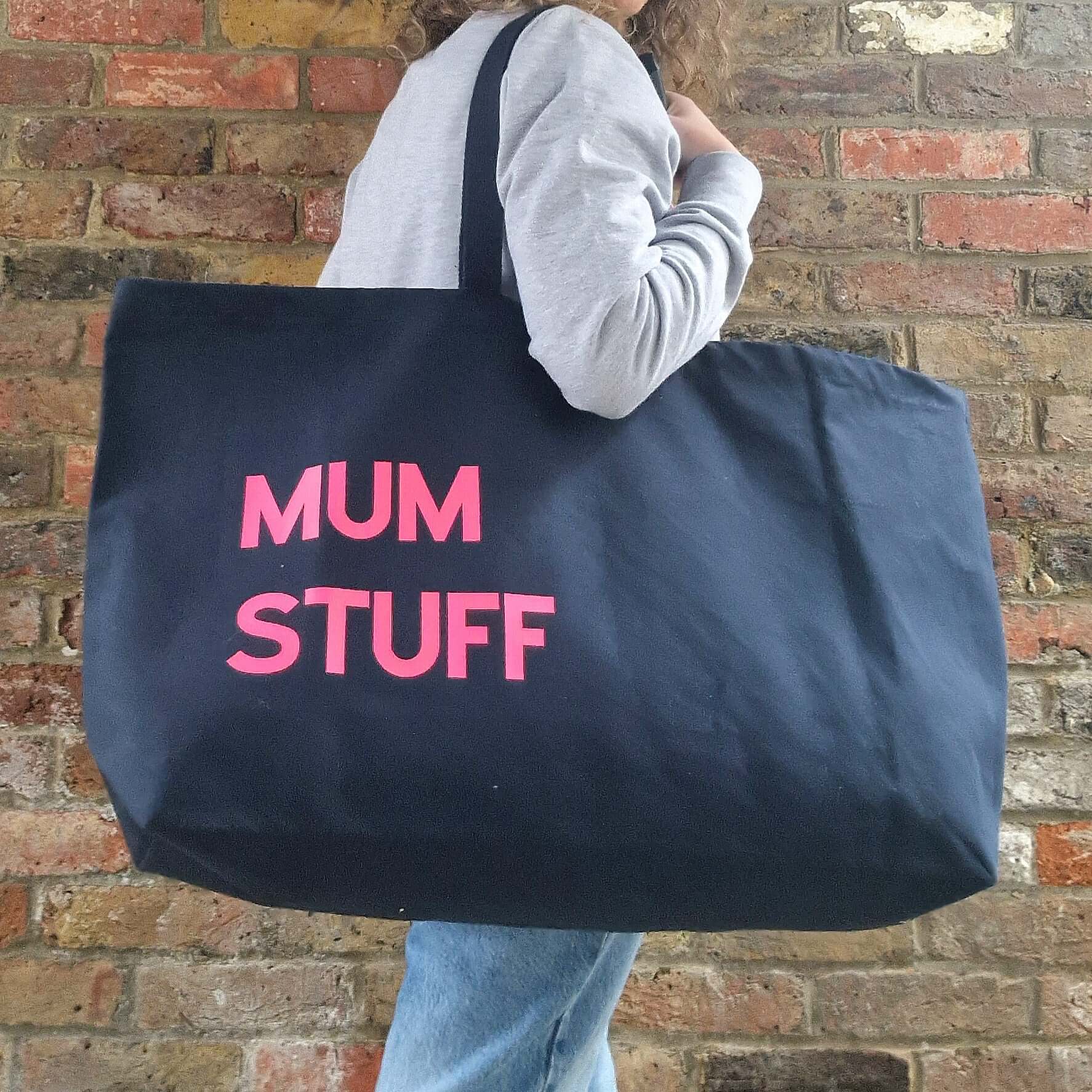 A person carrying a large black tote bag with the text 'MUM STUFF' printed in pink on the side, walking against a brick wall background.