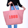 Pink tote bag with '1986' printed on it held by a person wearing blue jeans.