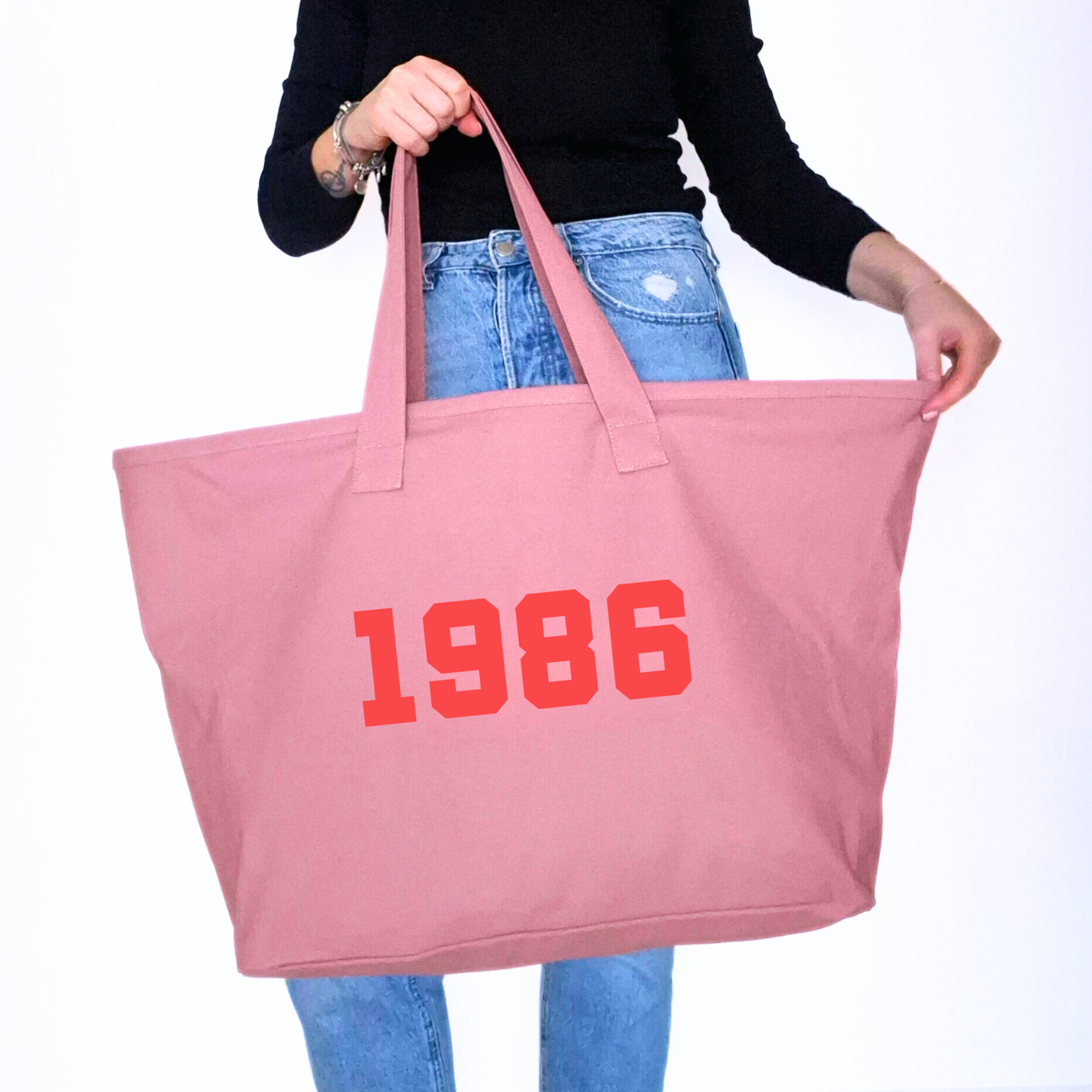 Pink tote bag with '1986' printed on it held by a person wearing blue jeans.