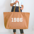 Person holding a tan tote bag with  personalised year '1986' printed on it against a plain background