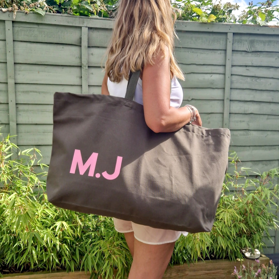 Person holding a brown tote bag with 'M.J.' initials in pink letters outdoors.