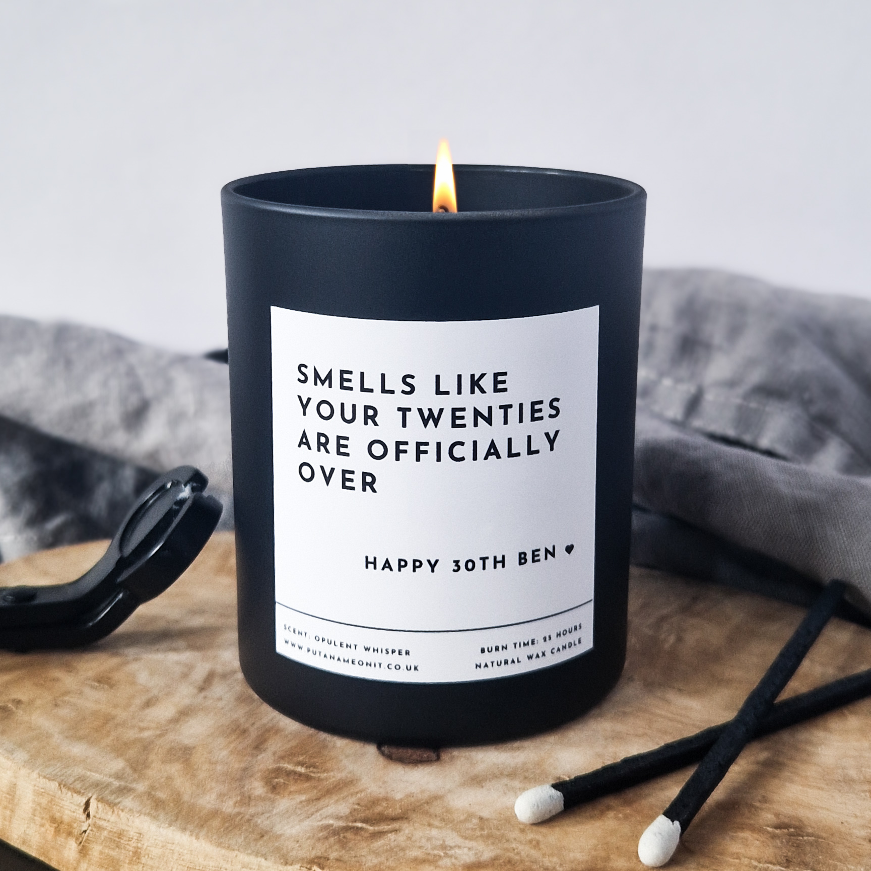 Black candle with a humorous label 'Smells like your  twenties are officially over' on a wooden surface
