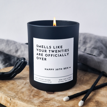 Black candle with a humorous label 'Smells like your  twenties are officially over' on a wooden surface