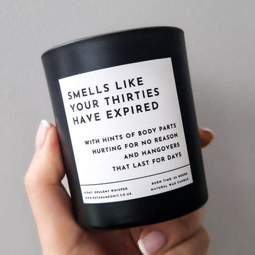 Matte Black candle with a white label that says 'Smells like you thirties have expired. With hint of body parts hurting for no reason and hangovers that last for days'