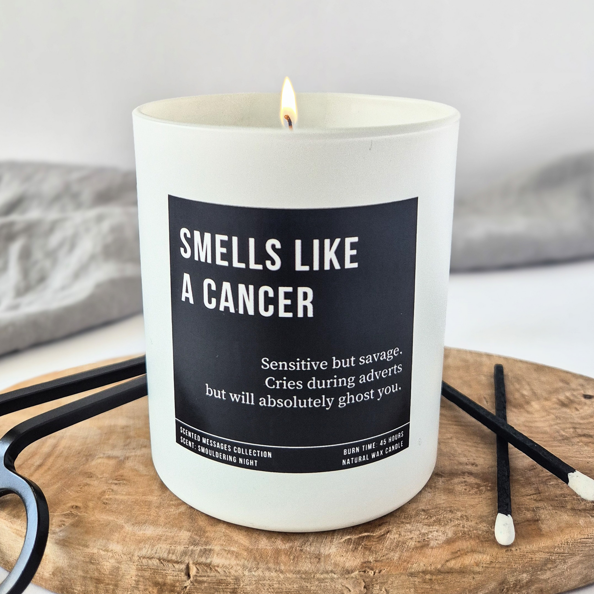 White candle with “Smells Like a Cancer” label and personalised traits below, styled with black matches and clean, minimalist background.