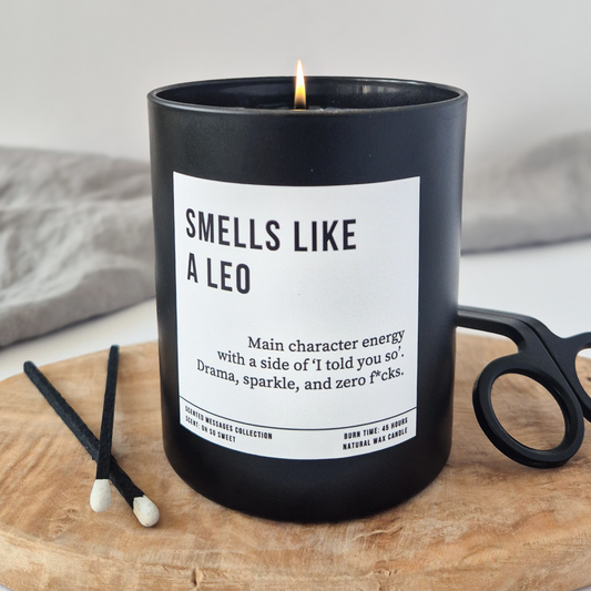 Black candle jar with white label that reads “Smells Like a Leo – Main character energy with a side of ‘I told you so’. Drama, sparkle, and zero fcks.” Styled on a wooden board with black candle snuffer and matches.