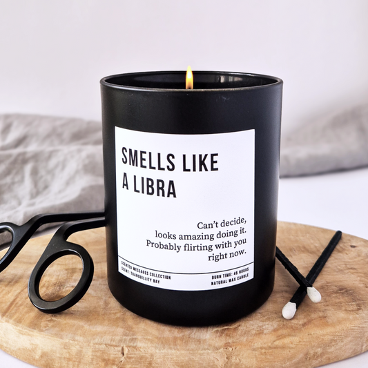 Black candle with a label on a wooden board with scissors and matches. With text 'smells like a Libra'