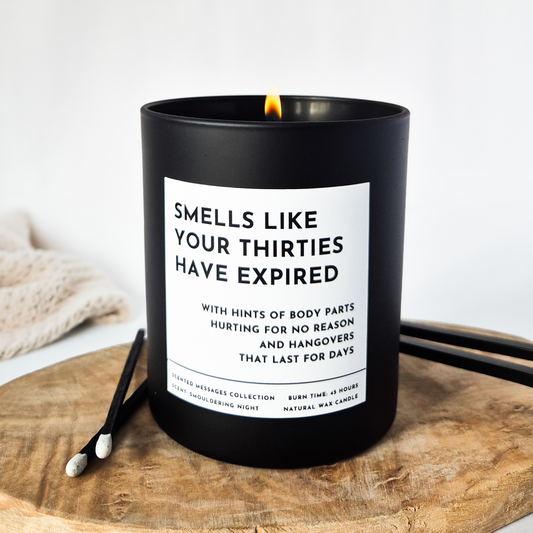 Black candle with a humorous label 'Smells like your thirties have expired' on a wooden surface