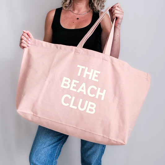 Oversized pale pink tote bag with customisable slogan ‘The Beach Club’, held in hand.