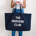 Person holding a navy tote bag with 'The Weekend Club' text on a plain background