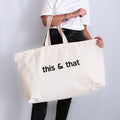Oversized natural canvas tote bag with customisable black slogan ‘this & that’, held in both hands.