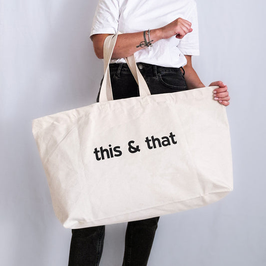 Oversized natural canvas tote bag with customisable black slogan ‘this & that’, held in both hands.