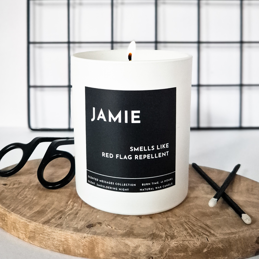 White candle with a black label and a text with personalised name and a text ' smells like a red flag repellent'  on a wooden surface.