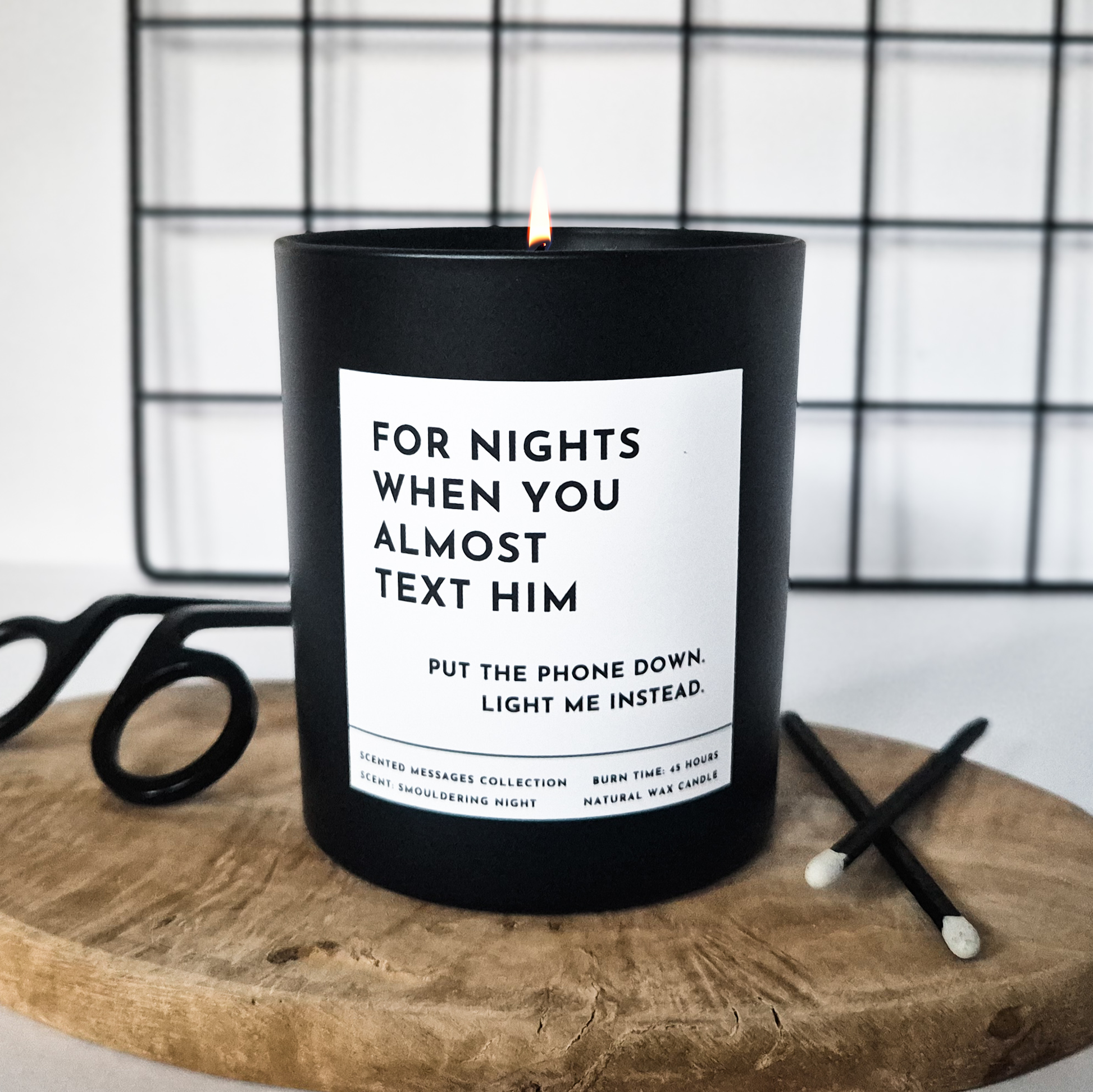 Black candle with a humorous label 'For the night when you almost text him. Put the phone down and light me instead' on a wooden surface