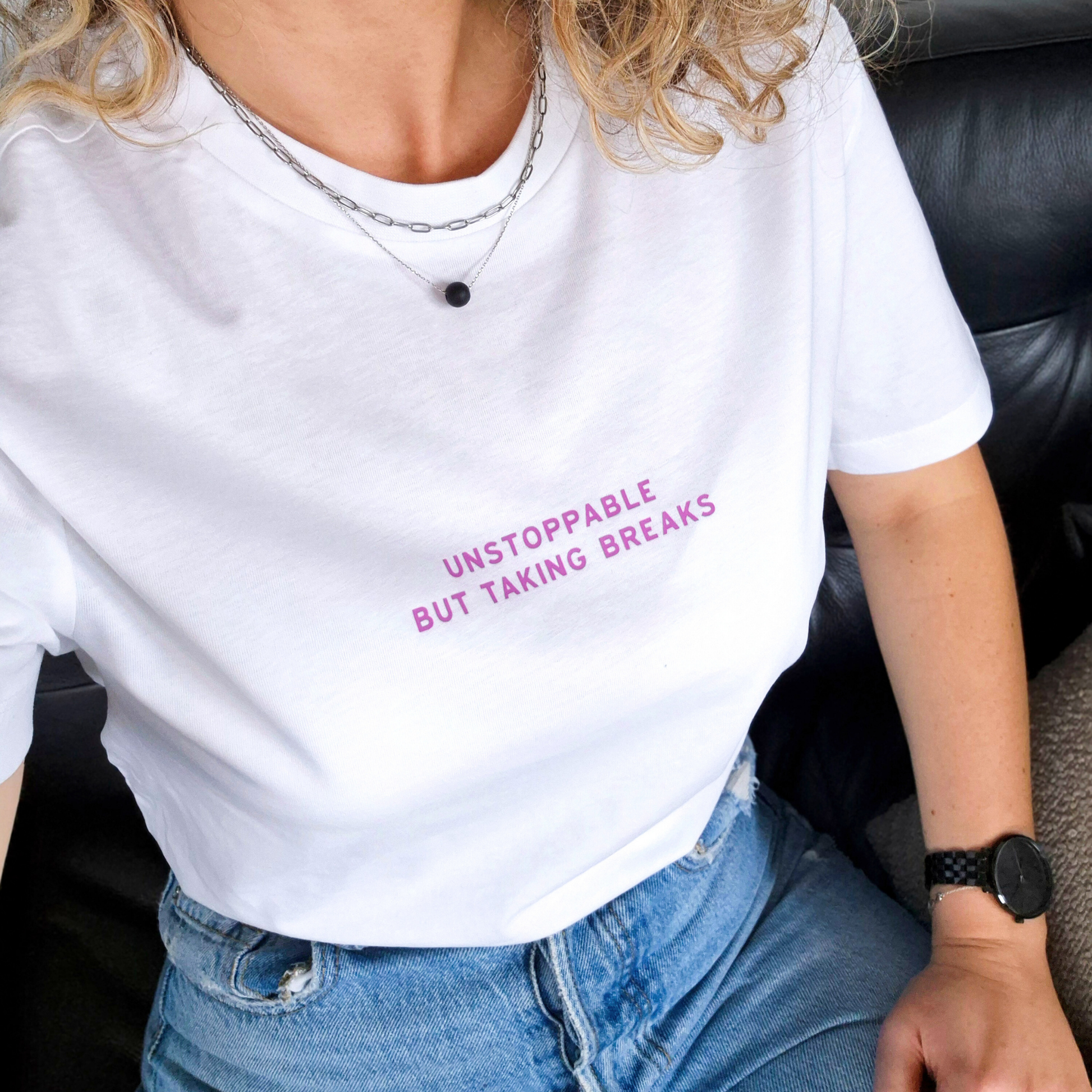 Person wearing a white t-shirt with pink text 'Unstoppable but taking breaks' sitting on a black leather couch.