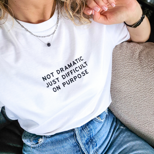 Person wearing a white t-shirt with text 'NOT DRAMATIC JUST DIFFICULT ON PURPOSE' sitting on a couch.