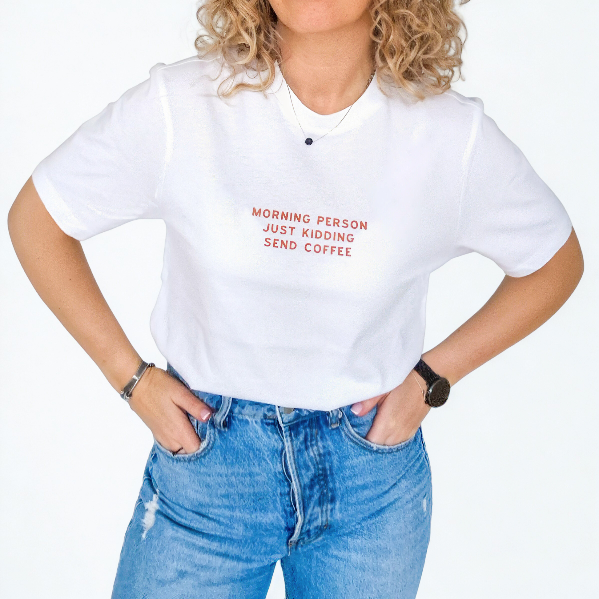 Person wearing a white t-shirt with text 'Morning person just kidding send coffee' and blue jeans on a plain background