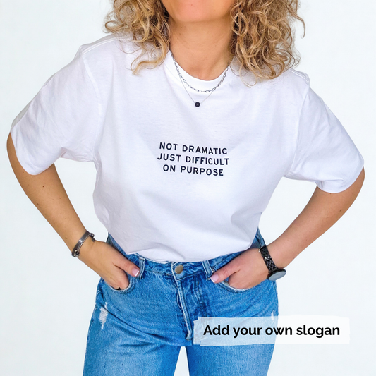 Person wearing a white t-shirt with a printed slogan 'Not dramatic just difficult on purpose' and blue jeans on a light grey background