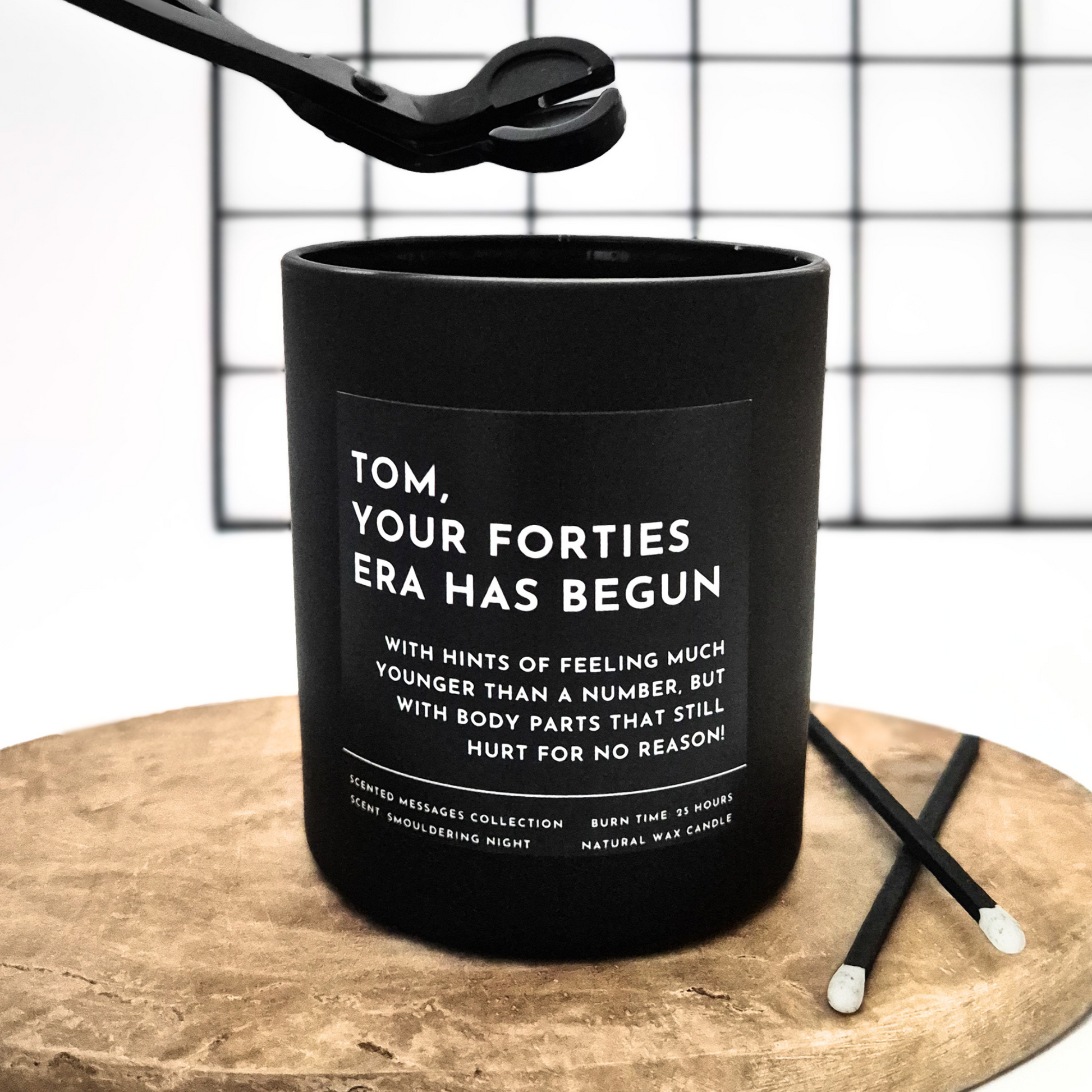 Black candle with text 'Your forties era has begun' and a personalised message on a wooden surface and grid background