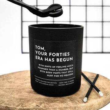 Black candle with text 'Your forties era has begun' and a personalised message on a wooden surface and grid background