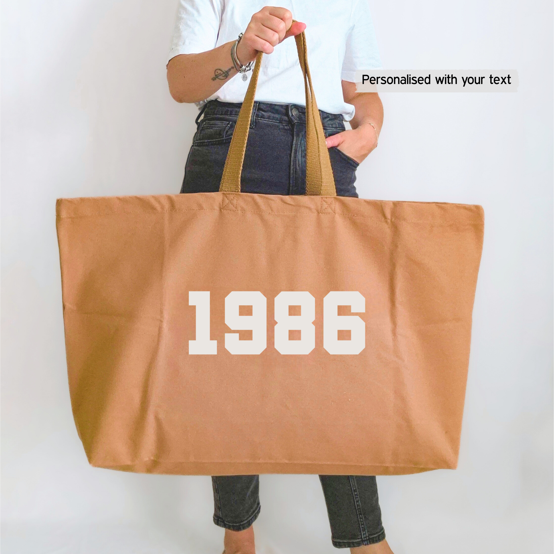 Person holding a tan tote bag with '1986' custom year printed on it against a plain background
