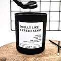 Black candle with a white label and text 'Smells like new start' on a wooden surface and grid background