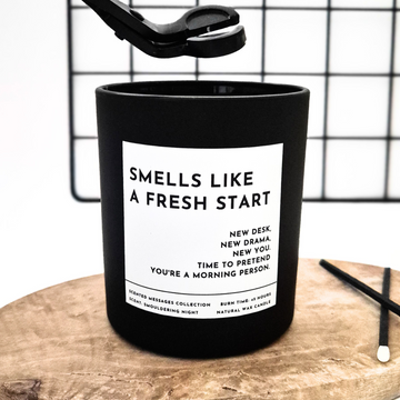 Black candle with a white label and text 'Smells like new start' on a wooden surface and grid background