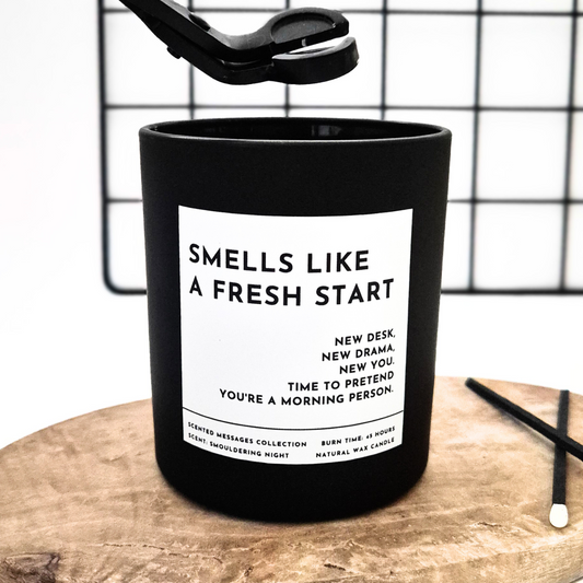 Black candle with a white label and text 'Smells like new start' on a wooden surface and grid background