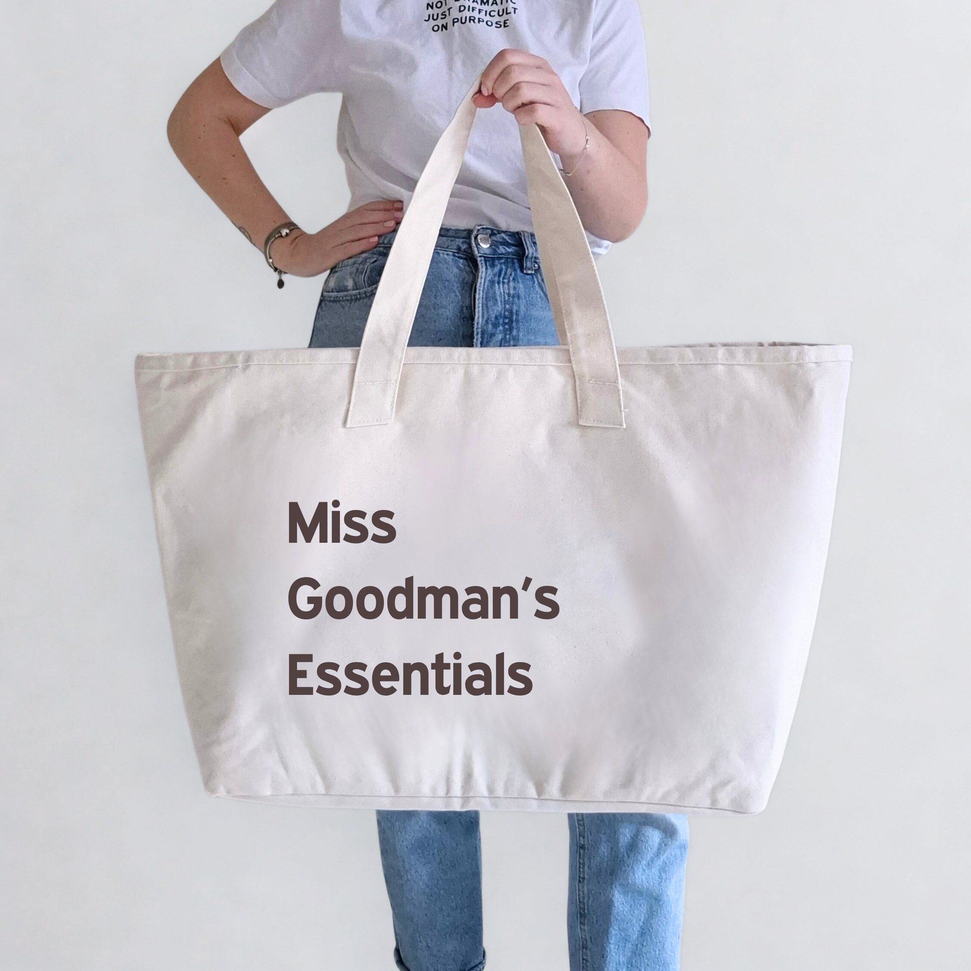 Person holding a beige tote bag with 'Miss Goodman's Essentials' text on a plain background. Text can be personalised