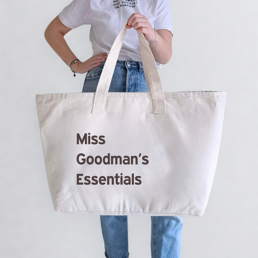Person holding a beige tote bag with 'Miss Goodman's Essentials' text on a plain background. Text can be personalised