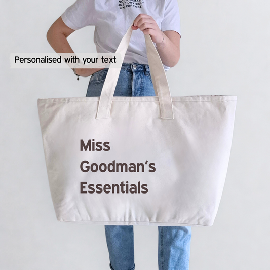 Person holding a beige tote bag with 'Miss Goodman's Essentials' text on a plain background. This photo shows that text can be personalised 