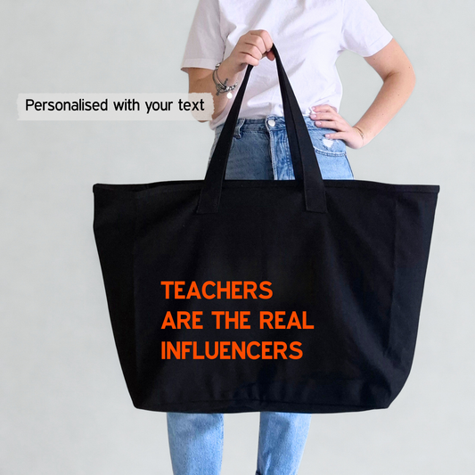 Person holding a black tote bag with 'Teachers are the real influencers' text in orange and a text on the photo that says 'Personalised with your text' so that customers can choose any text they wat