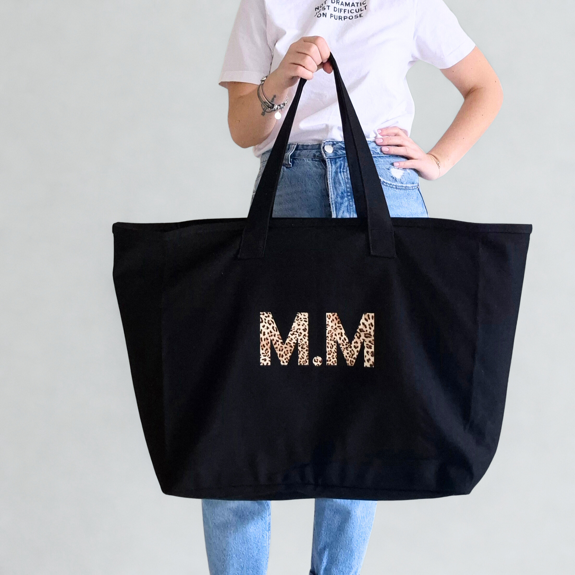 Person holding a black tote bag with 'M.M' in leopard print letters against a plain background