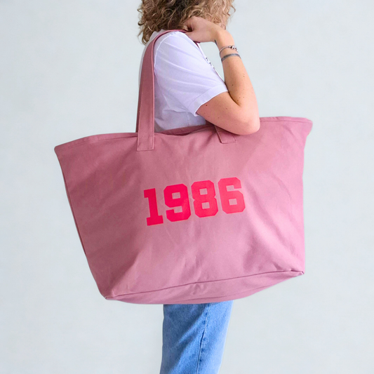 Person holding a pink tote bag with '1986' printed on it against a light gray background