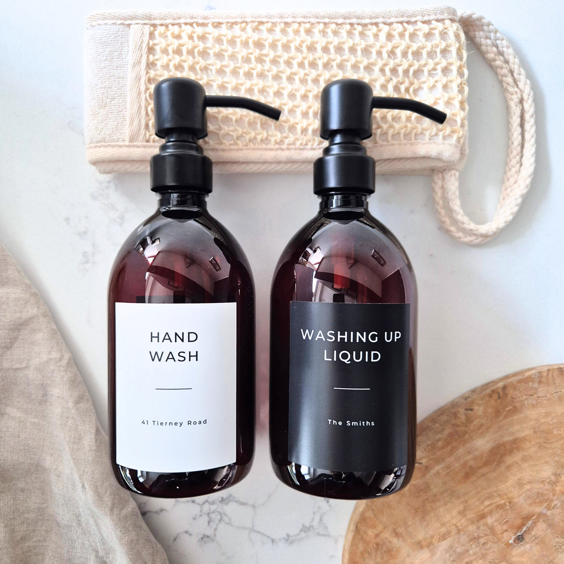 Two brown bottles labeled 'Hand Wash' and 'Washing Up Liquid' on a marble surface with a loofah in the background.