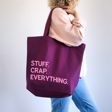 A person wearing a light pink sweatshirt and blue jeans carries a large deep purple canvas tote bag with pink text that reads 'STUFF. CRAP. EVERYTHING.