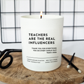 Candle with bold text 'Teachers are the real influencers' and smaller  message 'Thank you for everything you  do every single day. Year 6 2026'. on a wooden surface with a grid background