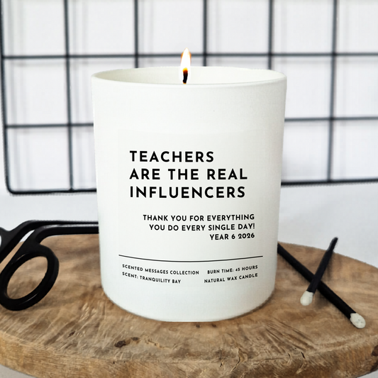 Candle with bold text 'Teachers are the real influencers' and smaller  message 'Thank you for everything you  do every single day. Year 6 2026'. on a wooden surface with a grid background
