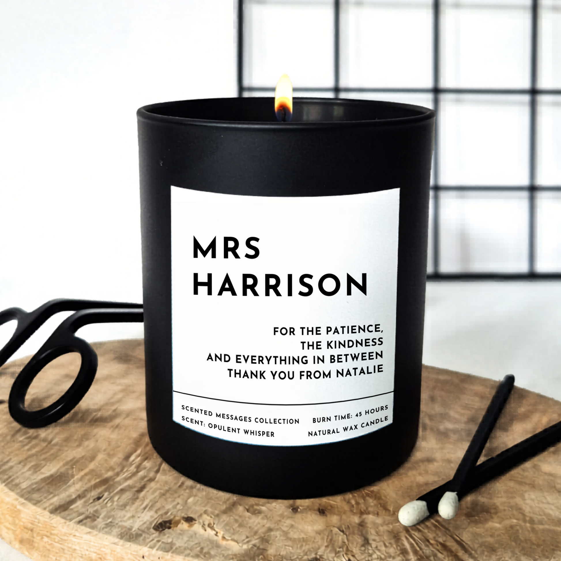 Black candle with a personalised label on a wooden surface. Text in bold 'Mrs Harrison' - a personalised teacher's name, and a personalised message underneath 'for the patience, the kindness and everything in between. Thank you from Natalie. 
